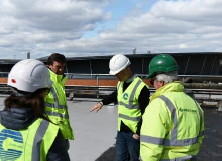 Group of four people wearing high visibility jackets and hard hats inspecting a flat roof, with a blue sky in the background