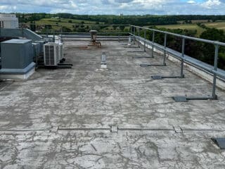 Weathered flat roof with peeling and deteriorated surface; air conditioning units and vents are positioned along the edge, overlooking green countryside.