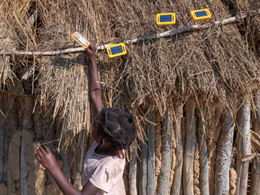 A young girl in a rural village hangs several portable solar lights to charge on the thatched roof of a hut.