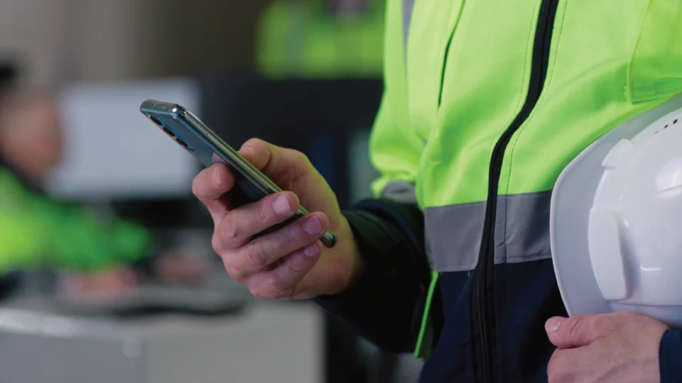 Construction worker in safety vest holding smartphone and hard hat outdoors