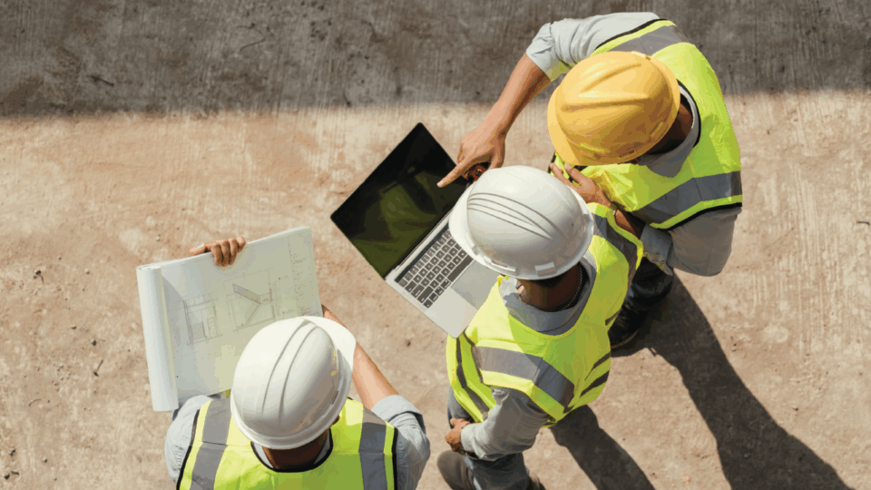 Three construction workers reviewing blueprints and laptop on site