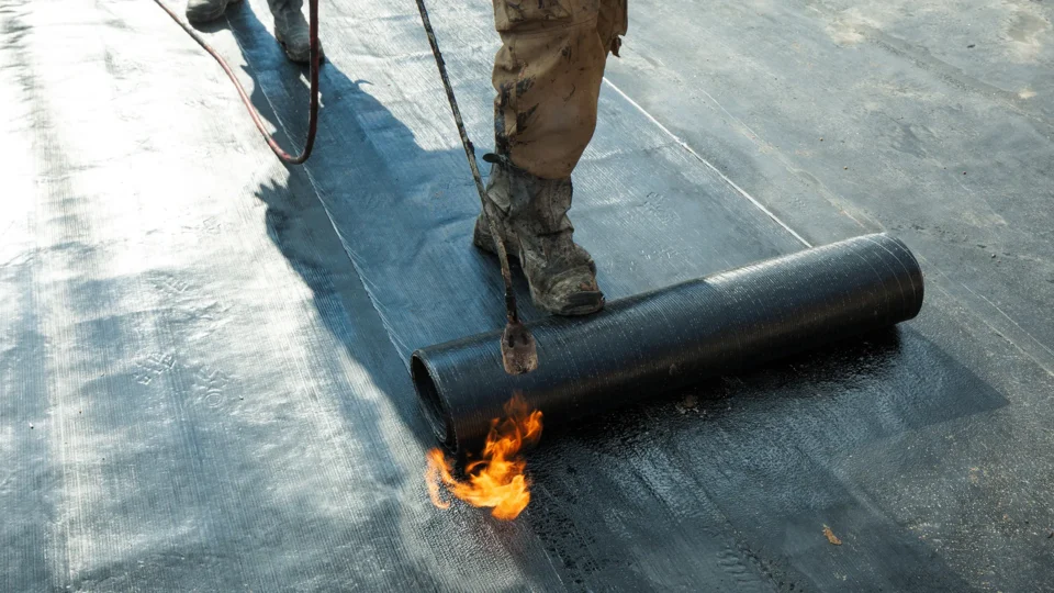 Worker using torch to seal bituminous roofing membrane on flat roof surface