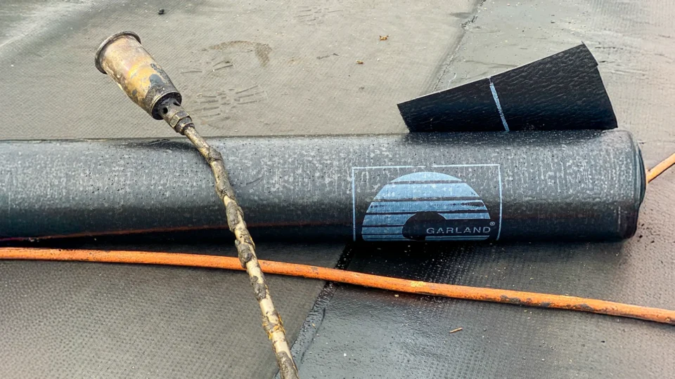 Roofing tools including torch, brush, and black membrane roll on wet surface