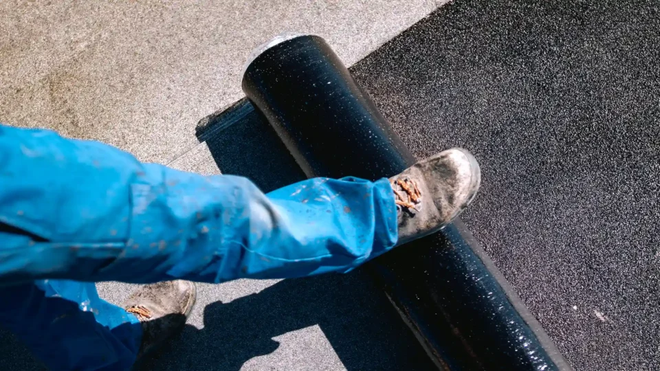 Worker in blue jacket applying black roofing membrane to asphalt surface