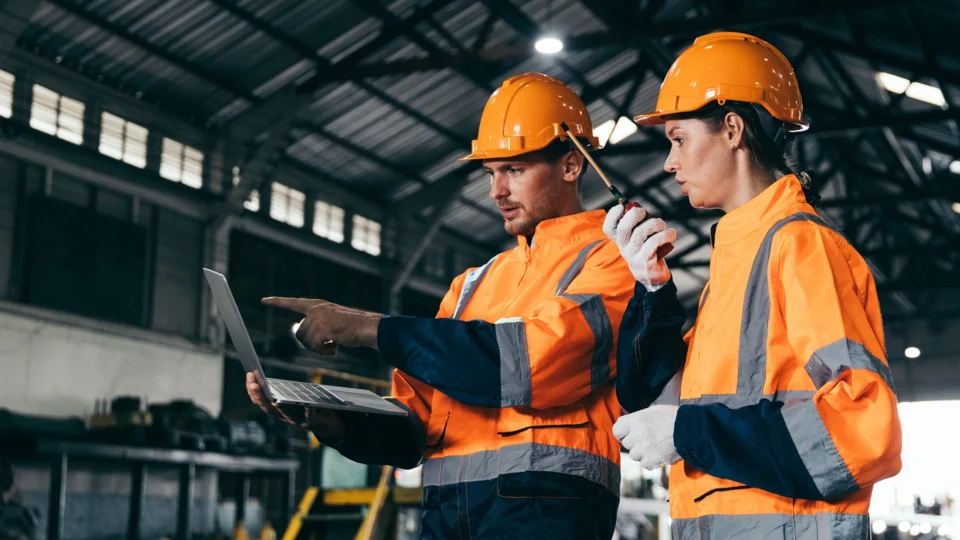 Two construction workers reviewing laptop data at industrial warehouse facility