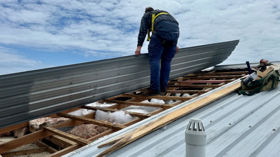 Worker inspecting metal roof with exposed wooden framing and insulation