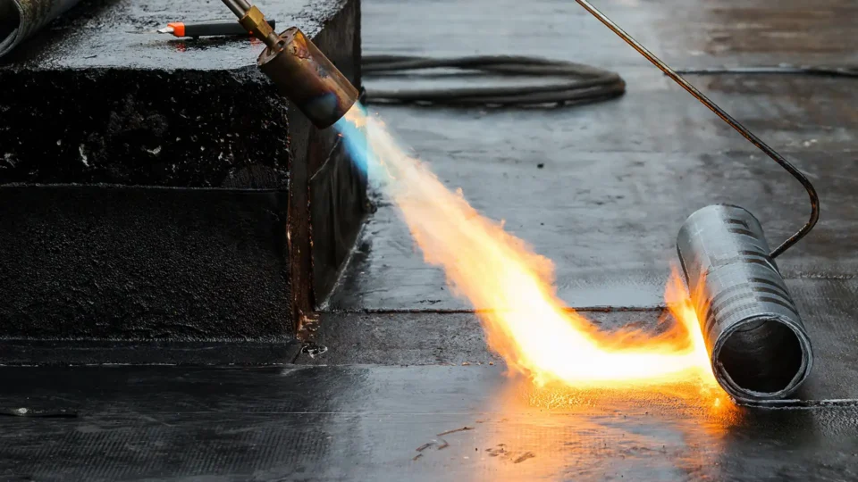 Blowtorch applying flame to roofing membrane installation on wet surface